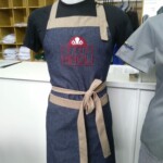 Avental frontal jeans leve com detalhes bege, bolsão e logo bordada na frente Avental com regulagem - Alfatex Uniformes Profissionais em Curitiba